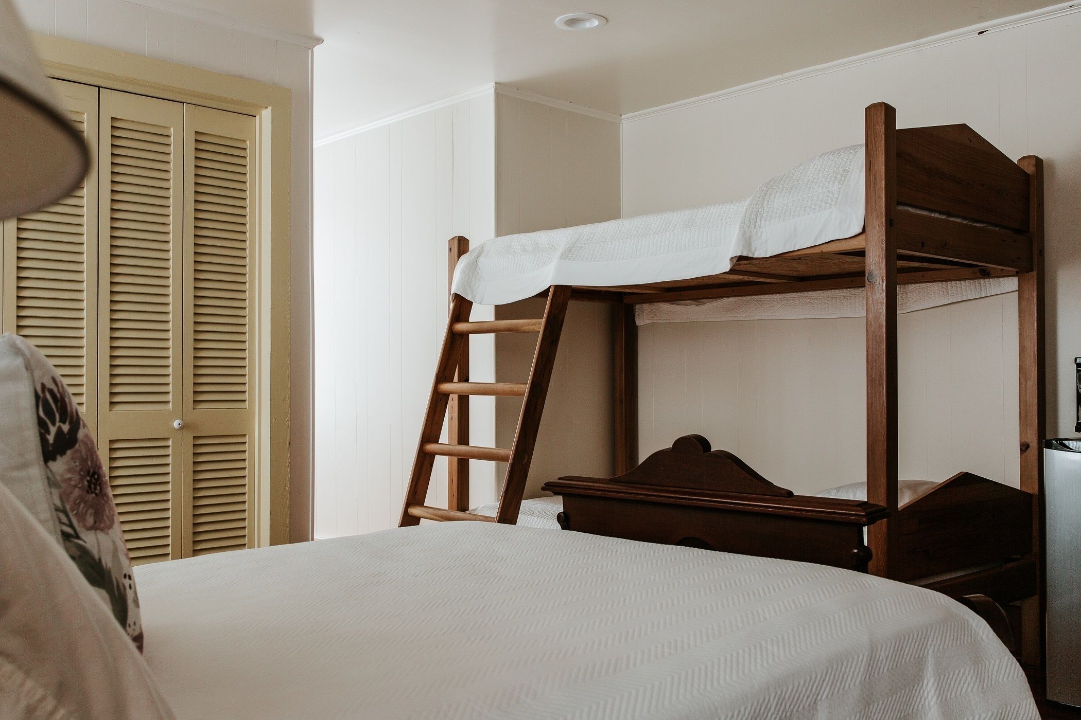 A bunk bed with a ladder, a white bedspread, and a wooden wardrobe with louvered doors.