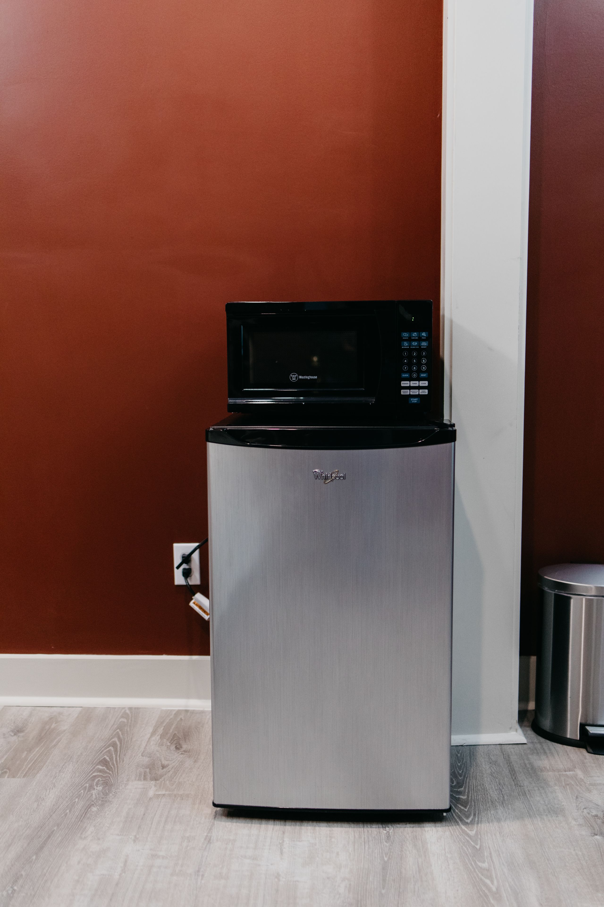 A compact refrigerator with a microwave on top, positioned against a red wall with a white baseboard.