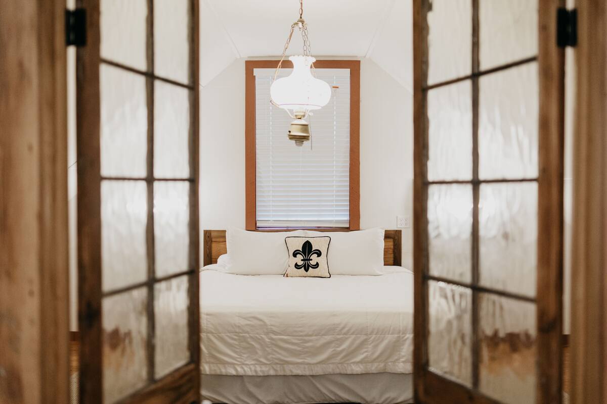 Une chambre à coucher avec un lit placé au centre, sous un luminaire suspendu, et de part et d’autre duquel se trouvent des portes en bois dotées de panneaux de verre.