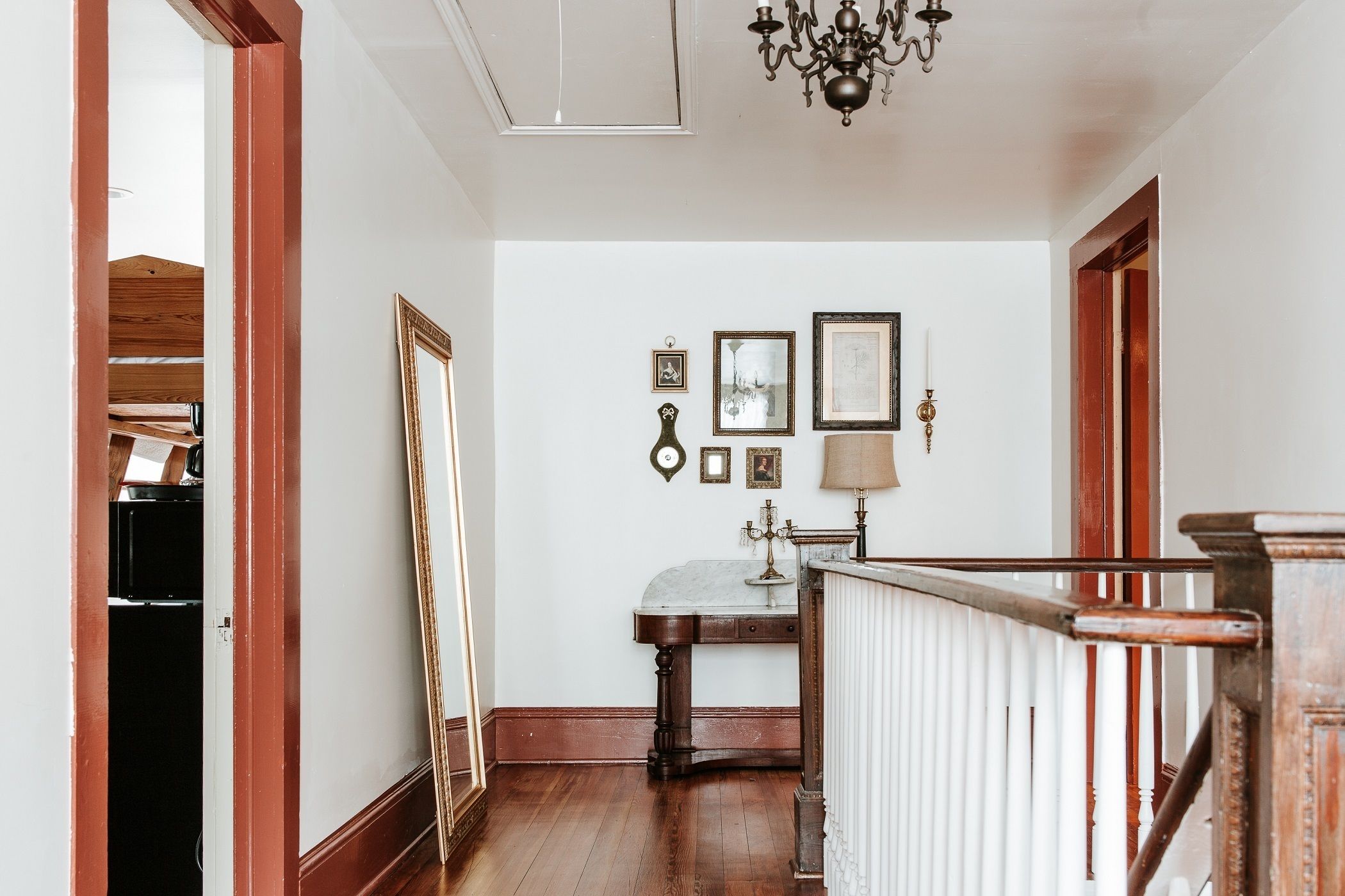 A hallway with a wooden floor, white walls, and a chandelier. There is a mirror leaning against the wall and a table with a lamp.