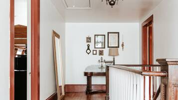 A hallway with a wooden floor, white walls, and a chandelier. There is a mirror leaning against the wall and a table with a lamp.