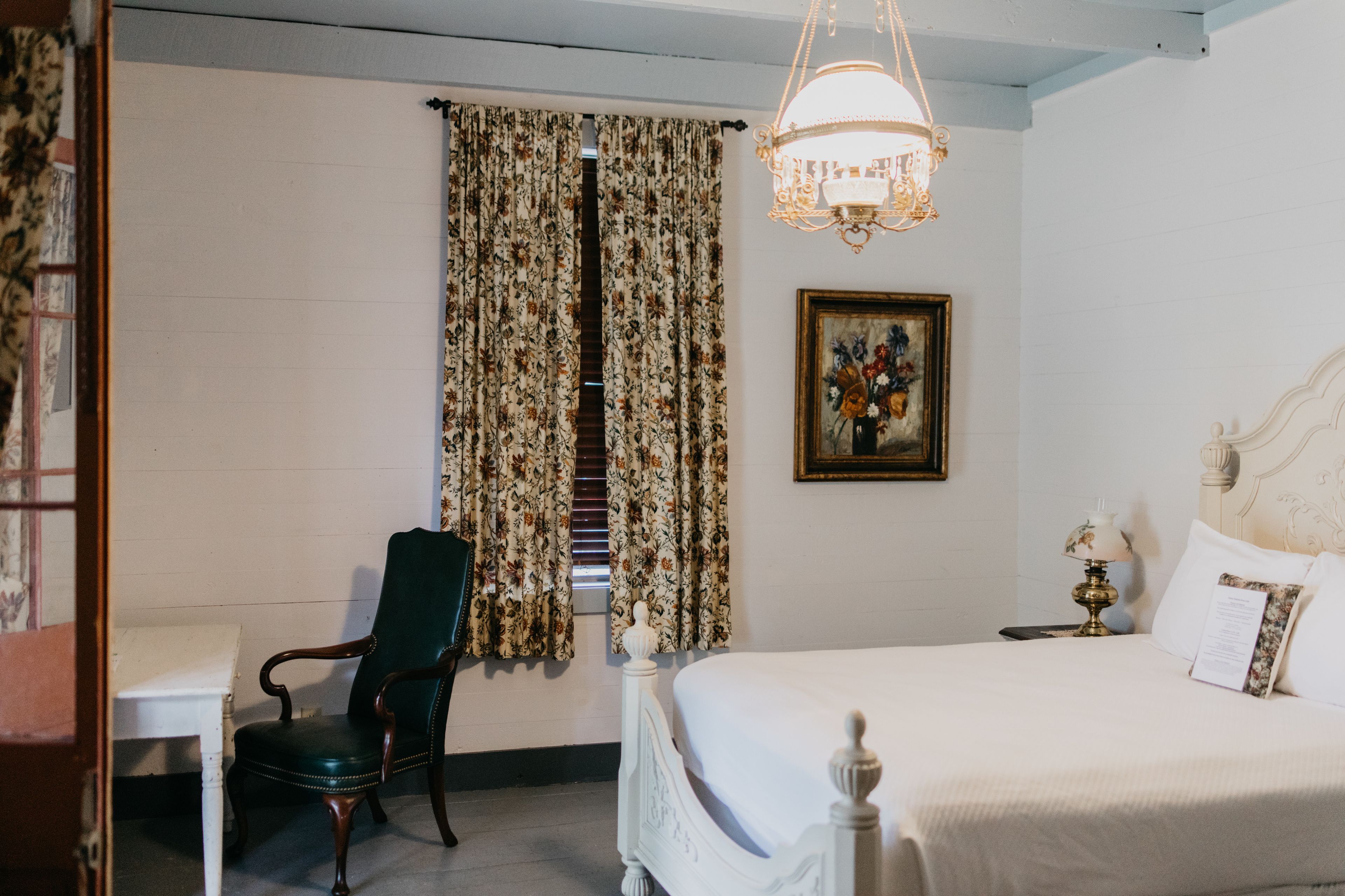 Une chambre d’hôtel avec un lit, une chaise, une table, une fenêtre avec des rideaux, un tableau et un lustre.