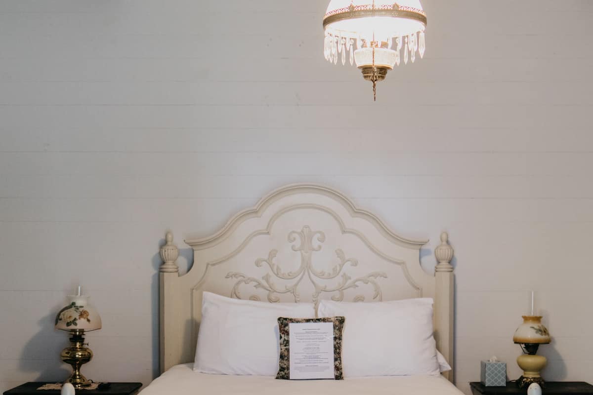 Un lit blanc orné, surmonté d’un lustre, deux tables de chevet avec des lampes et un mur sobre.