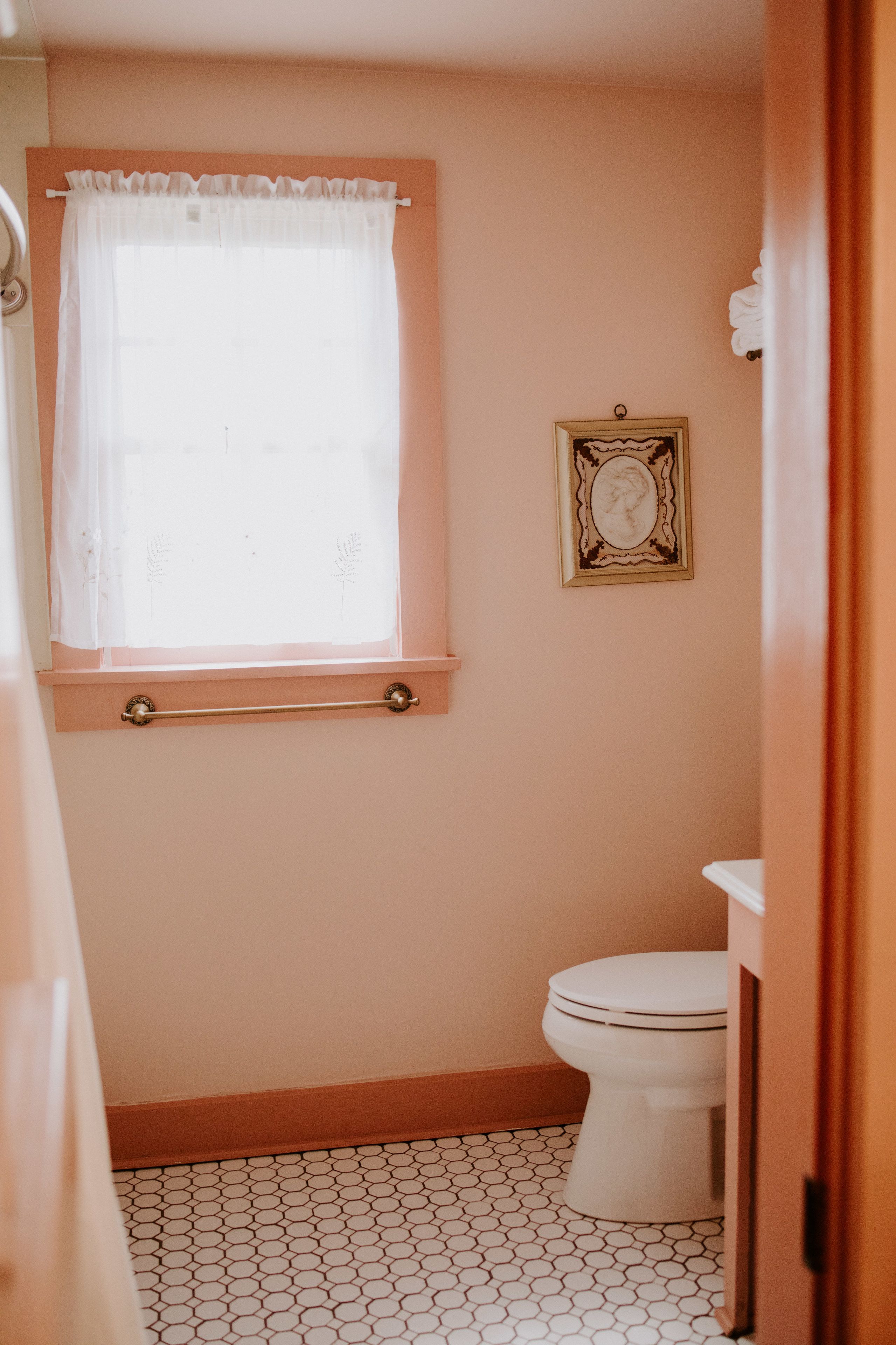A bathroom with a toilet, a window with a white curtain, a framed picture on the wall, and a tiled floor.