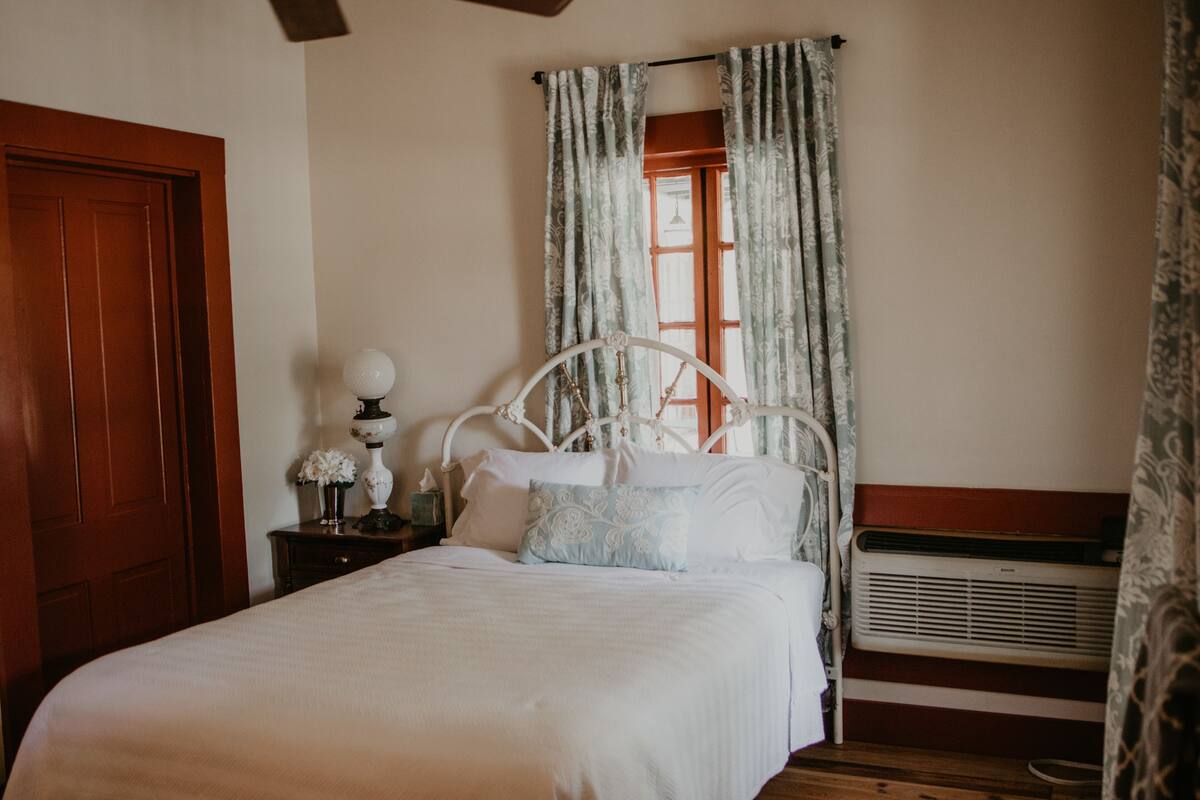 Une chambre avec une porte en bois, un lit blanc avec un cadre en métal, une table de chevet avec une lampe et une fenêtre avec des rideaux.