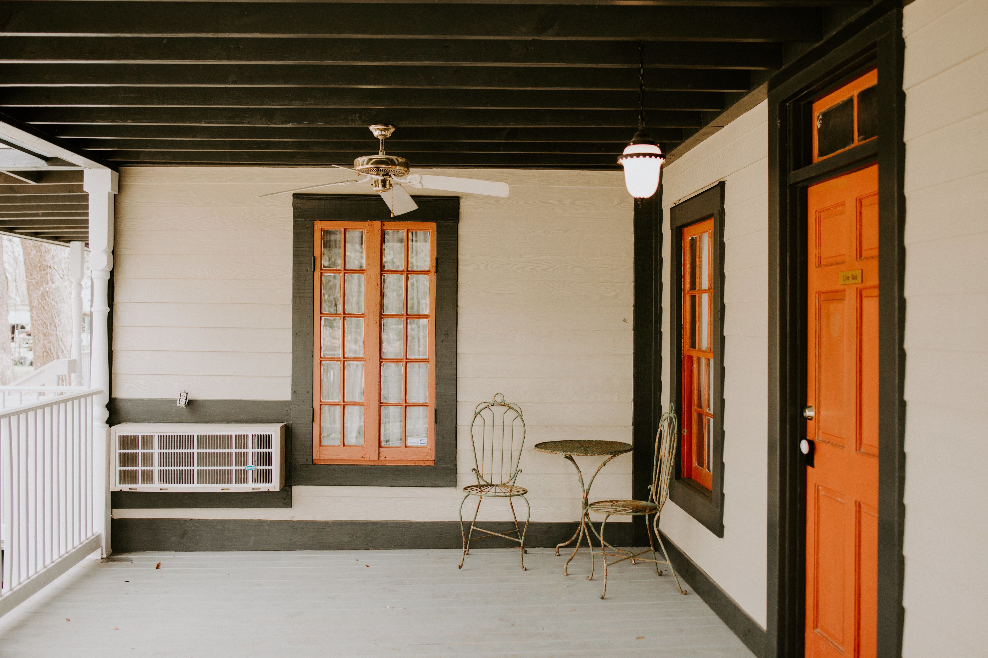 A covered porch with a ceiling fan, a window with a wooden frame, a metal chair, a small table, and an orange door.