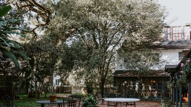 An outdoor patio with brick flooring, metal tables and chairs, potted plants, and a building with large windows in the background. - Mouton Plantation (Lafayette)