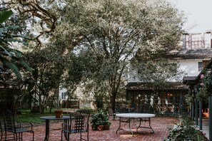 An outdoor patio with brick flooring, metal tables and chairs, potted plants, and a building with large windows in the background. - Mouton Plantation (Lafayette)