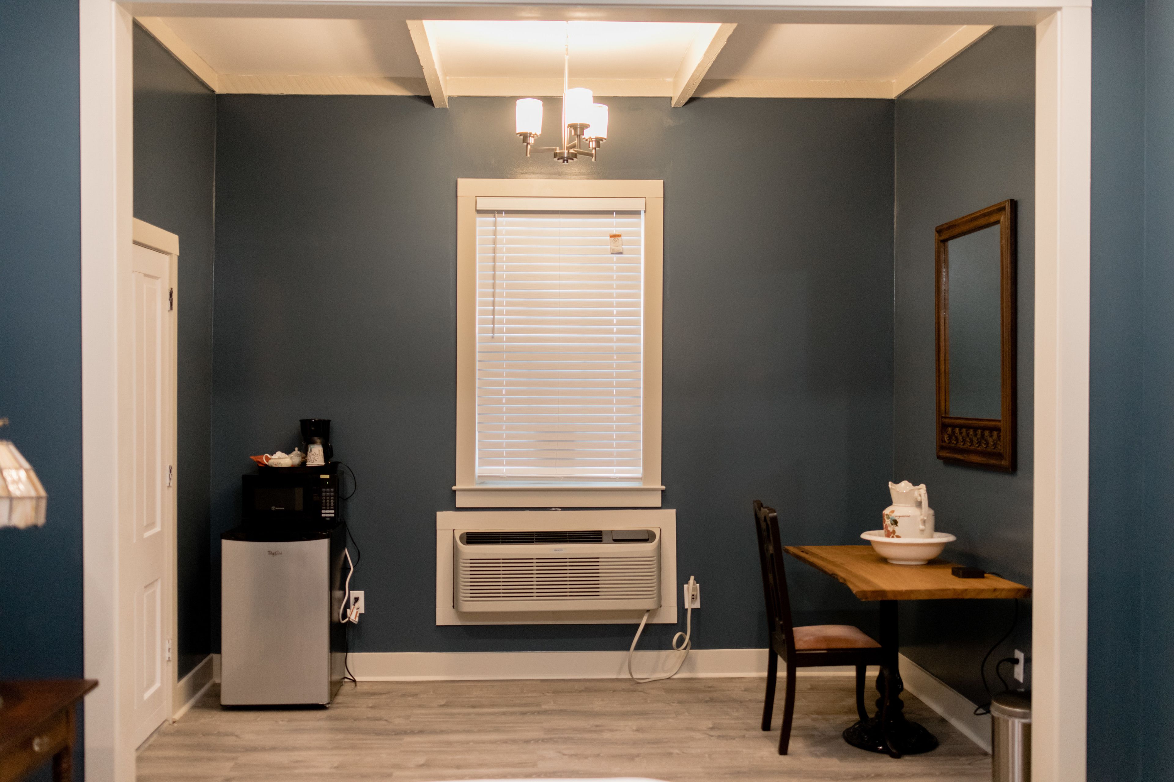 Una habitación compacta con aire acondicionado integrado, un refrigerador pequeño, una mesa de comedor con una silla y un espejo montado en la pared.