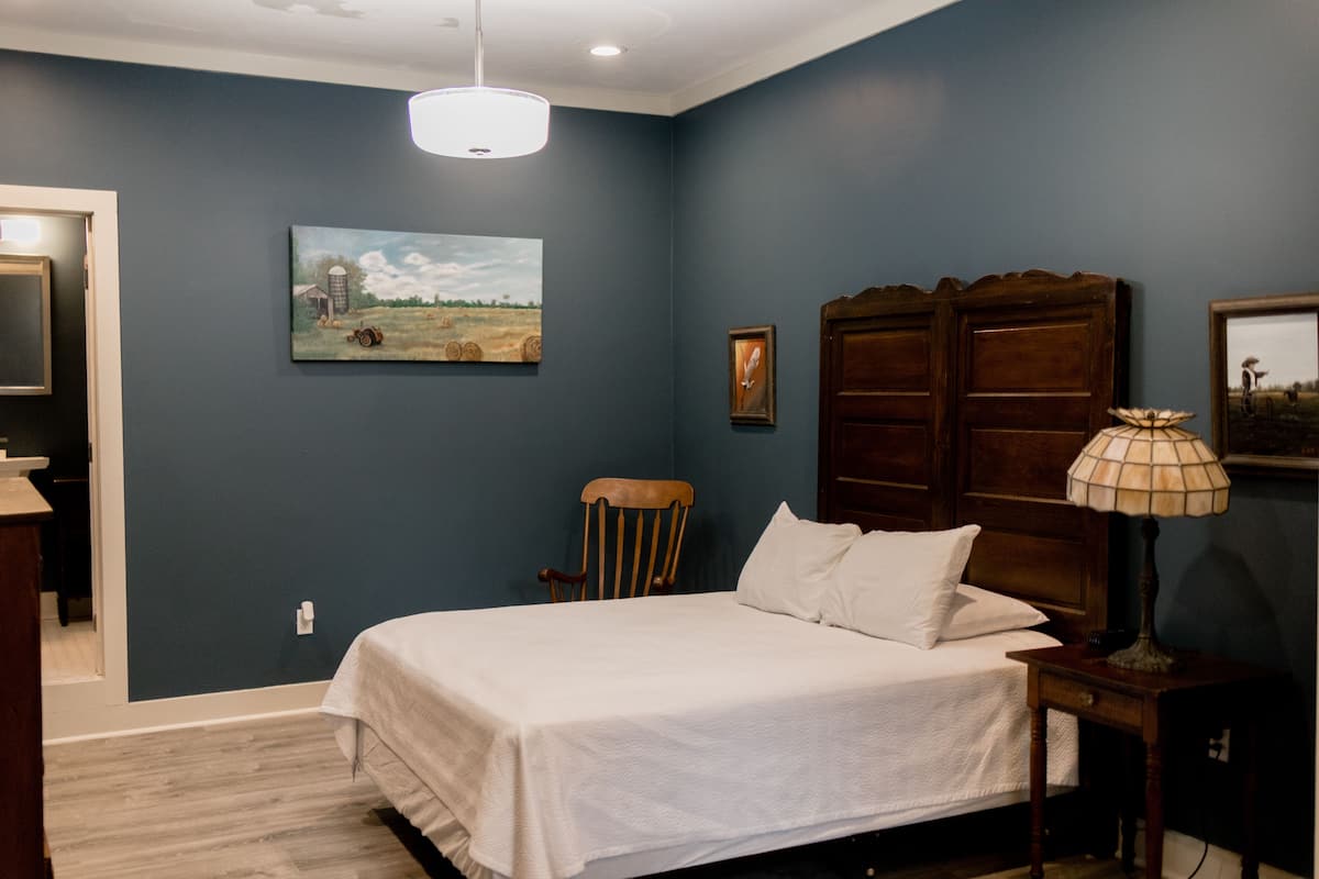 Une chambre à coucher avec un lit, une chaise en bois, une table de chevet, une lampe et un tableau accroché au mur.