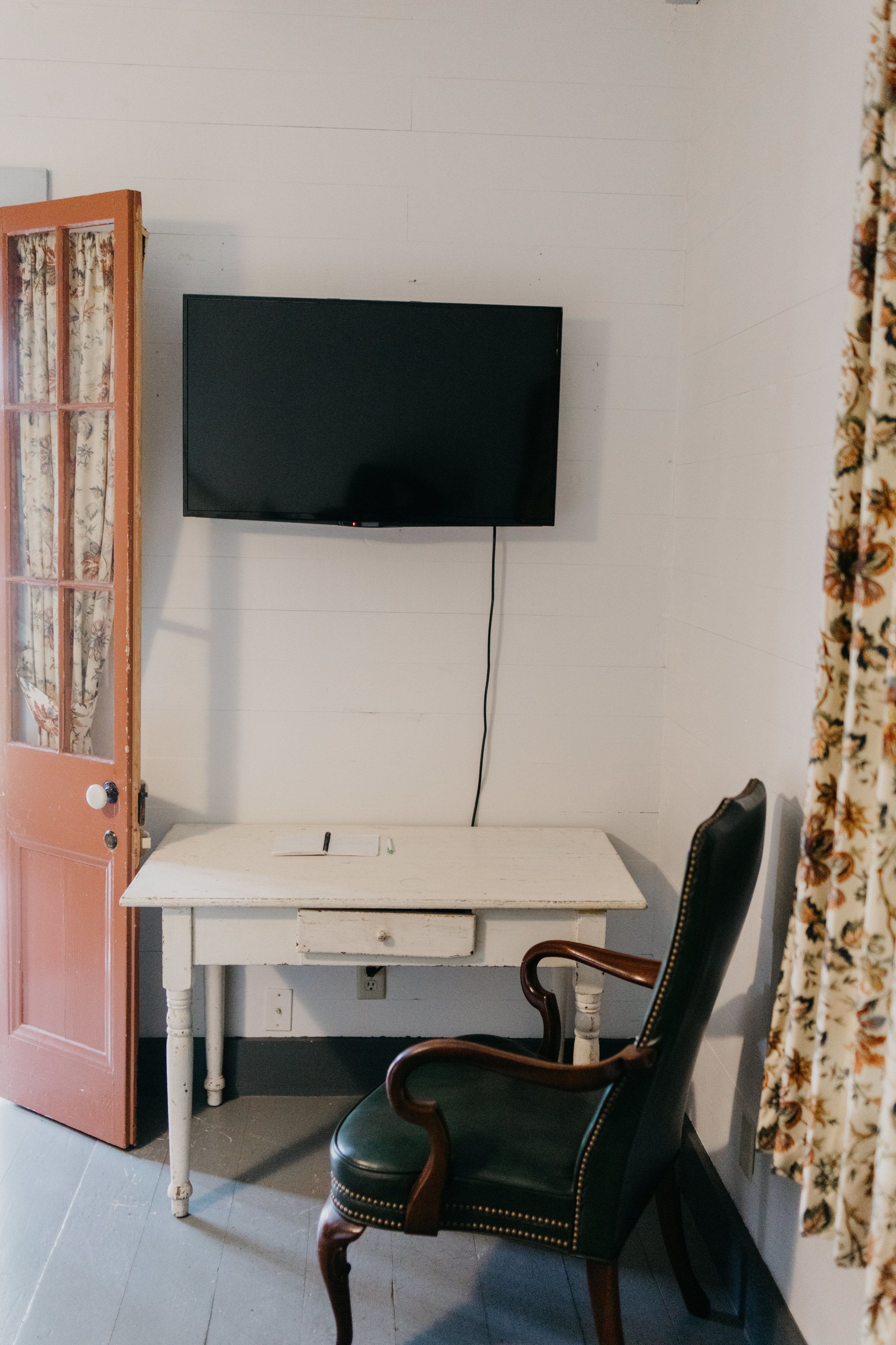 A room with a white desk, a wooden chair, a television mounted on the wall, and a door with floral curtains.