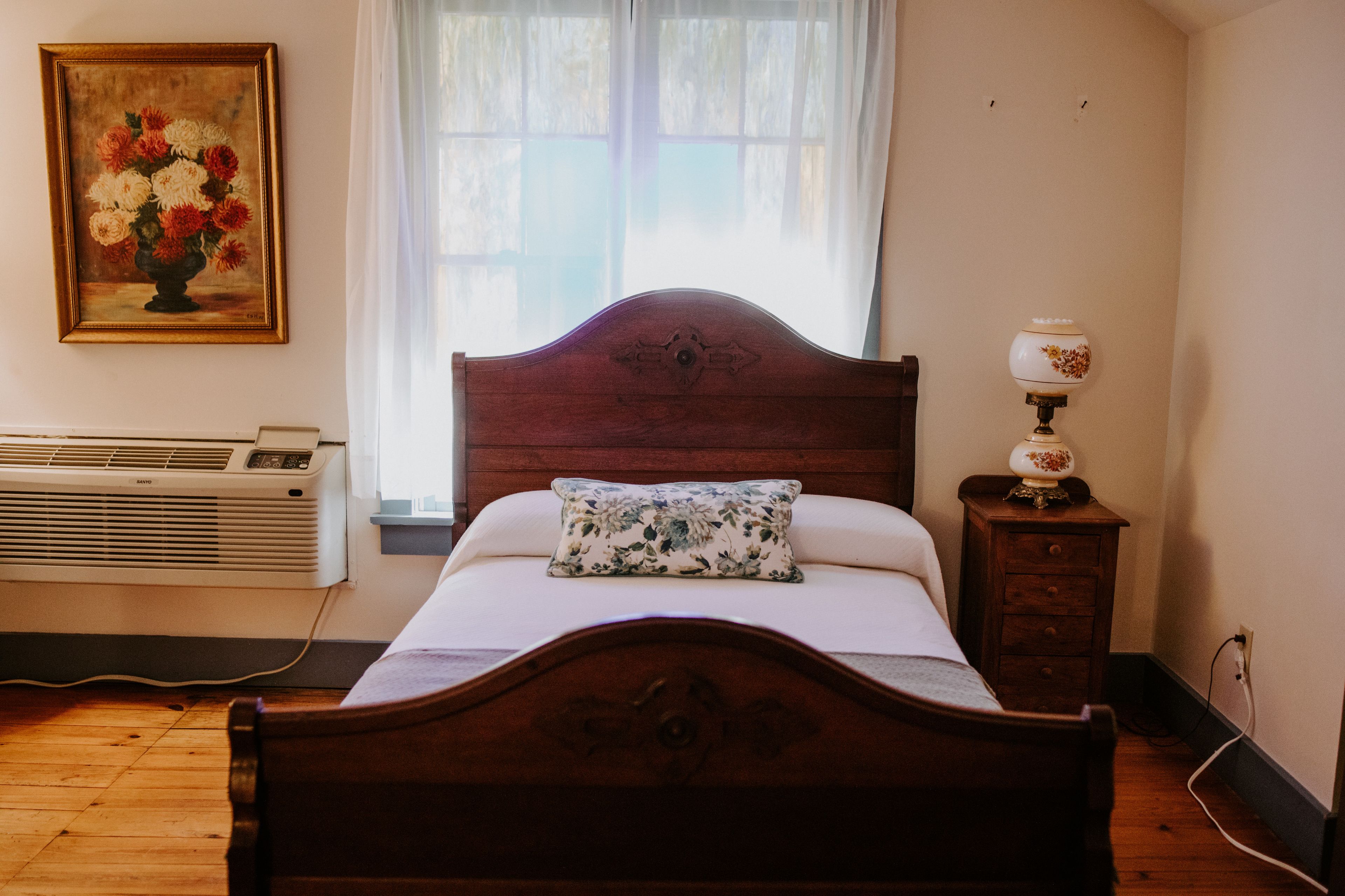 A bedroom with a wooden bed, a bedside table with a lamp, a radiator, and a window with curtains.