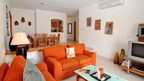 Ein modernes Wohnzimmer mit einem orangefarbenen Sofa, einem Essbereich mit Holzmöbeln und einem Fernseher auf einem Lowboard.