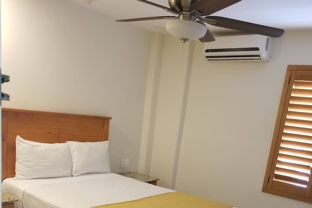 Una cama bien hecha con cabecera de madera, ventilador de techo y equipo de aire acondicionado en la pared.