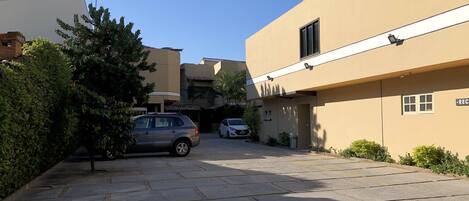 Un área de estacionamiento con dos coches, un edificio de fachada color beige y un árbol.