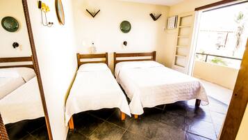 A room with two beds, a small table with a lamp, a wall-mounted shelf, and a window with a view of the outside.