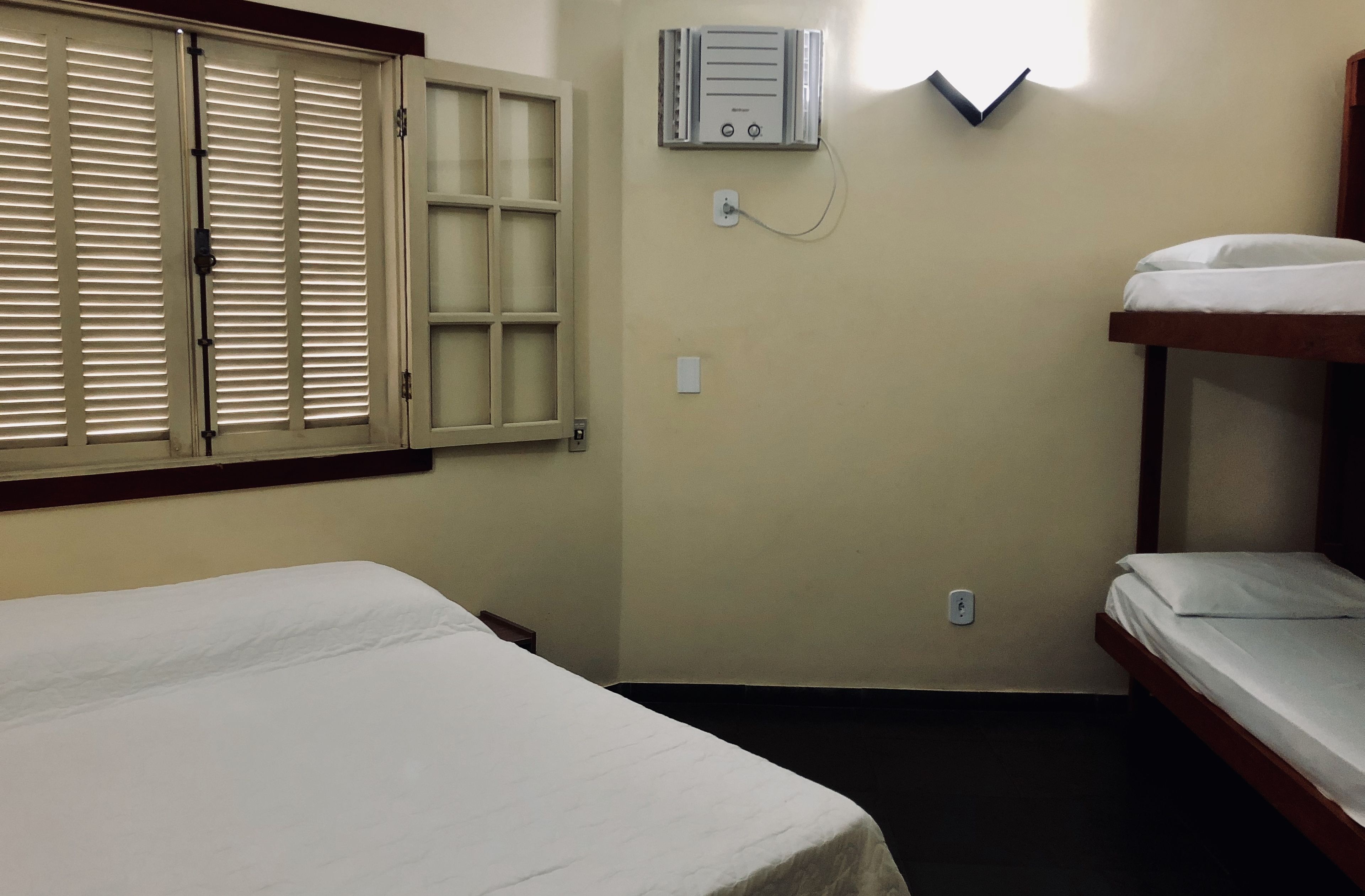 A small, simple room with two beds, a window with shutters, and a wall-mounted air conditioning unit.