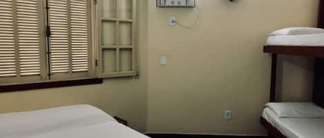 Una habitación pequeña y sencilla con dos camas, una ventana con contraventanas y un aire acondicionado de pared.