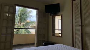 A room with a bed, a television mounted on the wall, a window with a view of palm trees, and a door with a mirror. - Pousada Gênesis (Arraial do Cabo)