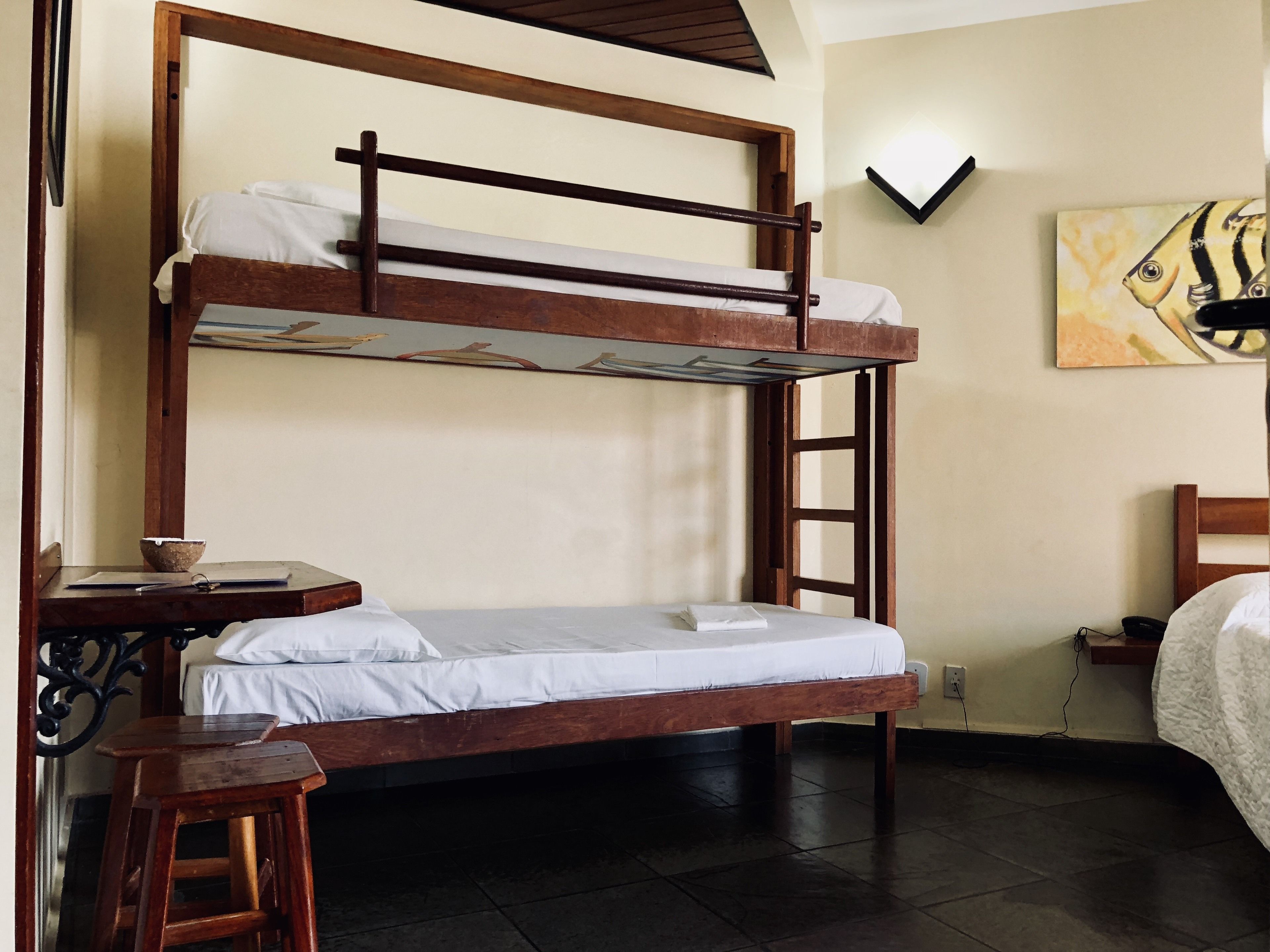 A bunk bed room with a desk and chair, a small table with a bowl, and a painting on the wall.