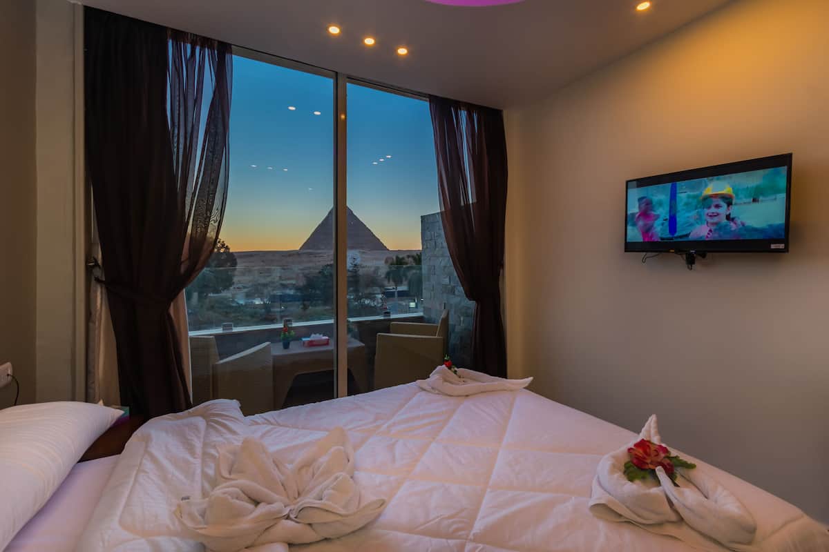Habitació d'hotel amb un llit gran, un televisor a la paret i vista d'una piràmide per la finestra.