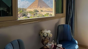 A room with a large window offering a view of a pyramid, two blue chairs, a small table with a vase of flowers, and a teapot.