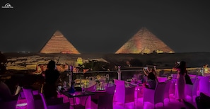 A rooftop bar with tables and chairs, a view of the pyramids, and a cityscape in the background.
