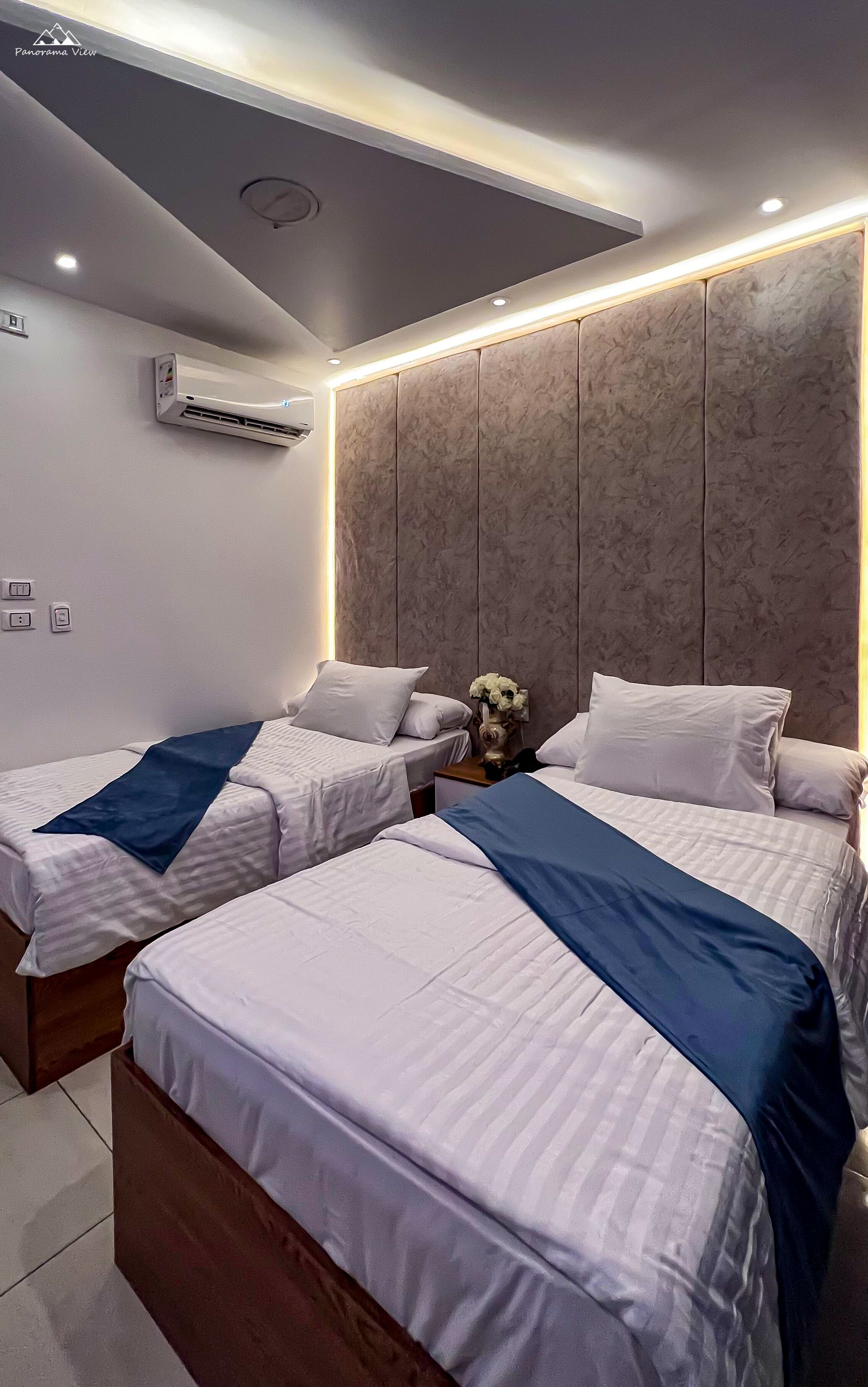 Una habitación de hotel con dos camas, cabecera de mármol, unidad de aire acondicionado y una mesita de noche con un jarrón de flores.