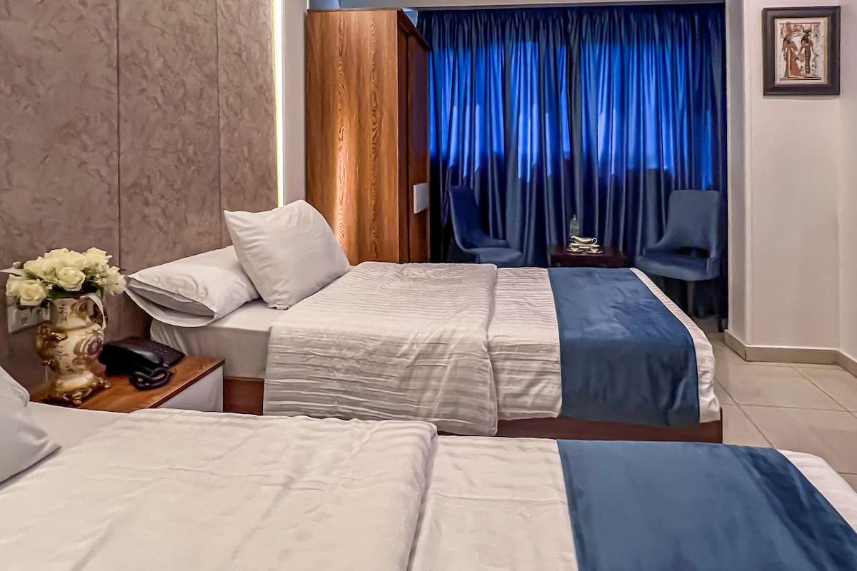 Habitació d'hotel amb dos llits, una taula de nit amb un telèfon i un ram de flors, una cortina blava i un quadre al mur.