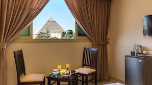 A hotel room with a view of a pyramid, a dining table with breakfast items, two chairs, and a minibar.