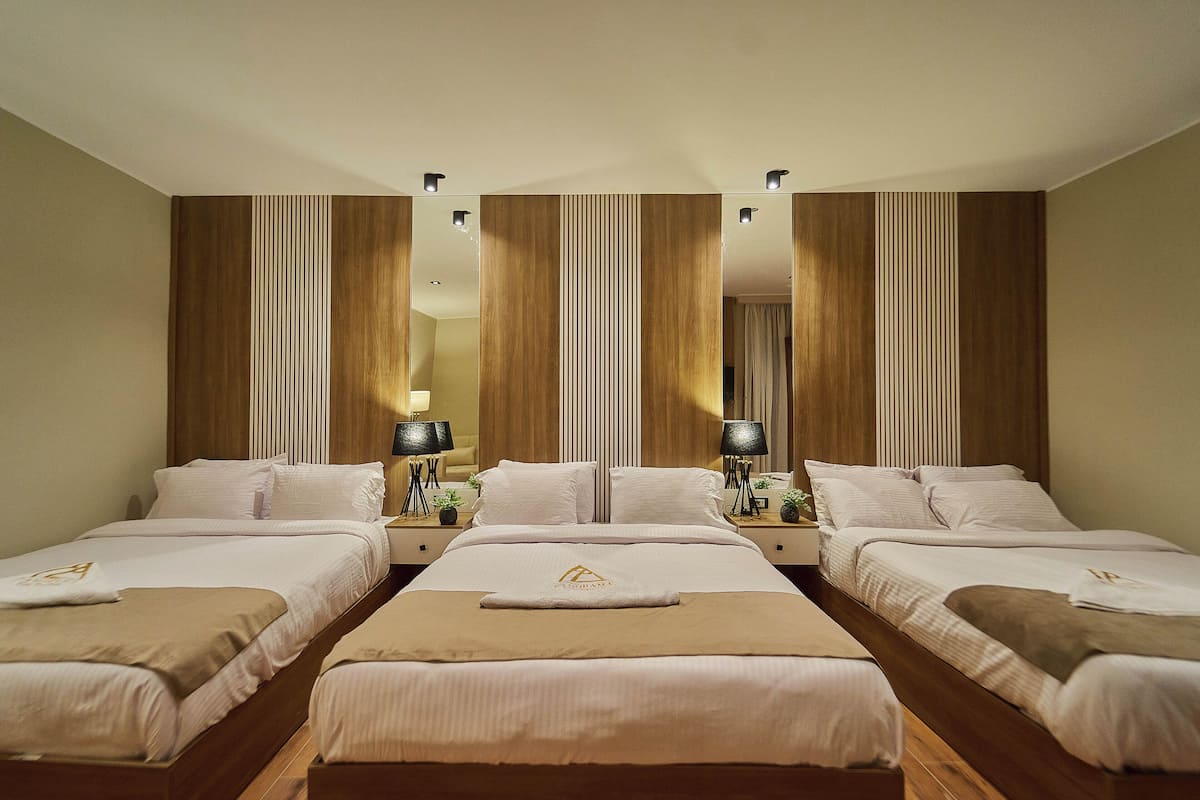 Una habitació d'hotel amb quatre llits alineats, cadascun amb roba de llit blanca i una manta marró. Hi ha dues taules de nit amb làmpades i una petita planta a cadascuna. La habitació té panells de fusta a les parets i un mirall gran sobre els llits.