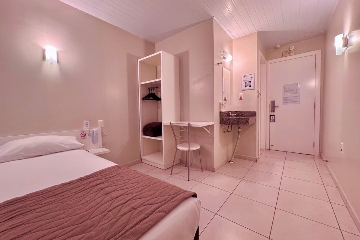 A hotel room with a bed, a desk, a chair, a bathroom with a sink, and a door.