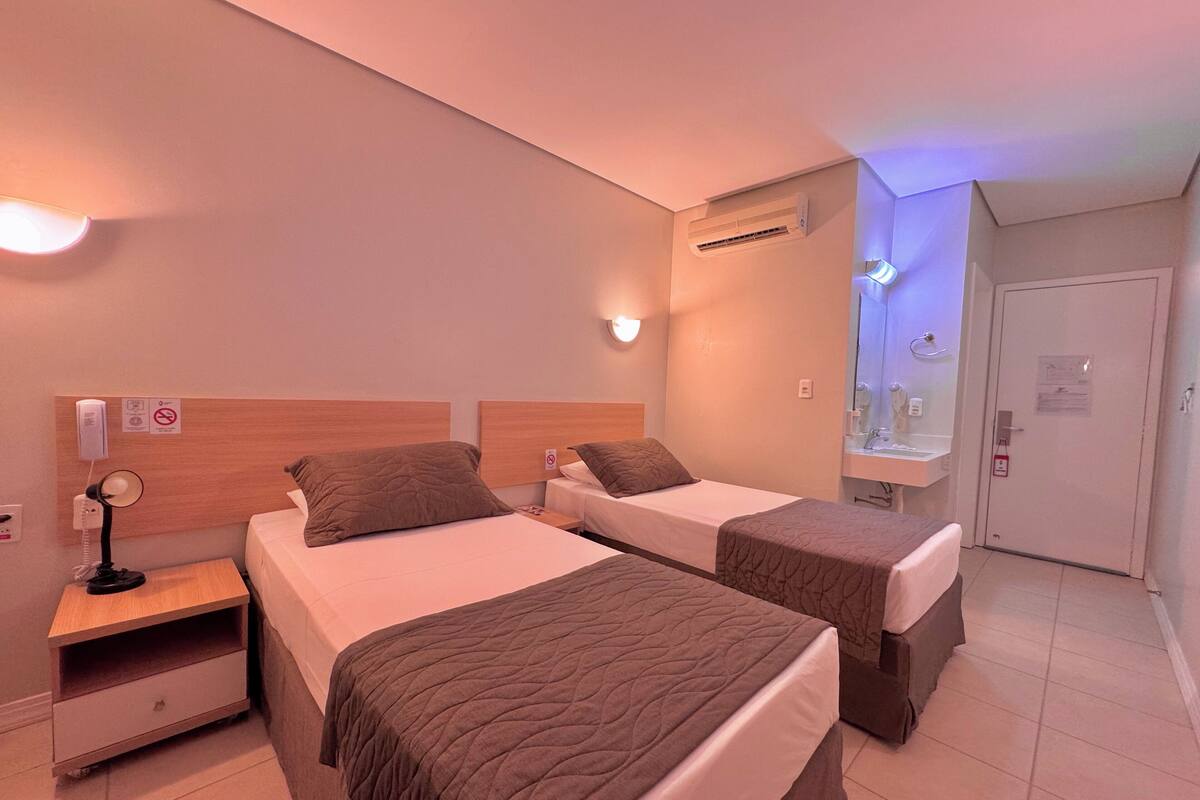 A hotel room with two beds, a bedside table with a lamp, an air conditioning unit, a mirror, and a door.