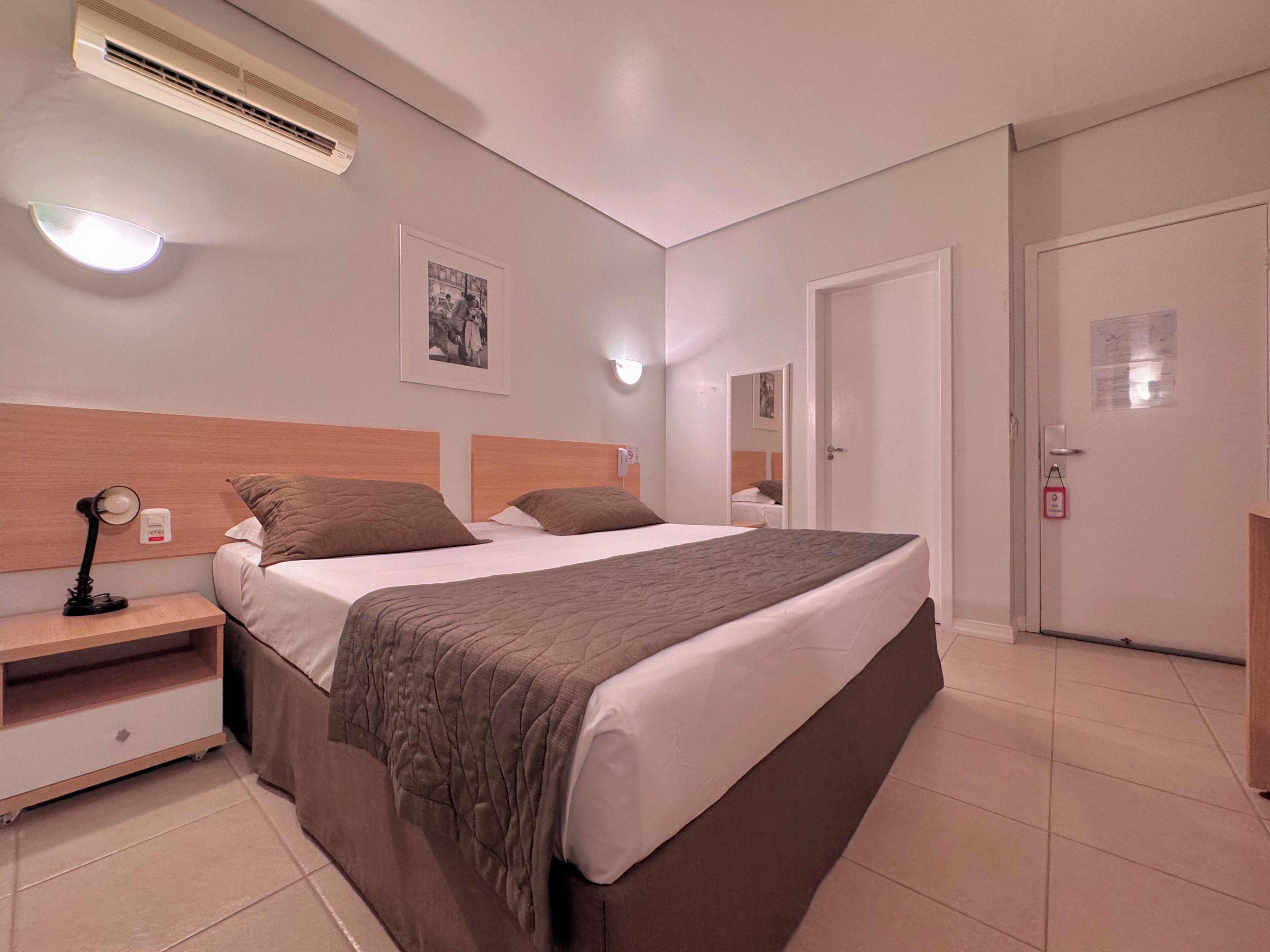 A hotel room with a large bed, bedside tables, a nightstand, an air conditioning unit, a wall-mounted light, and a framed picture.