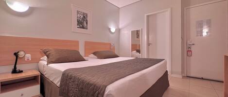 A hotel room with a large bed, bedside tables, a nightstand, an air conditioning unit, a wall-mounted light, and a framed picture.