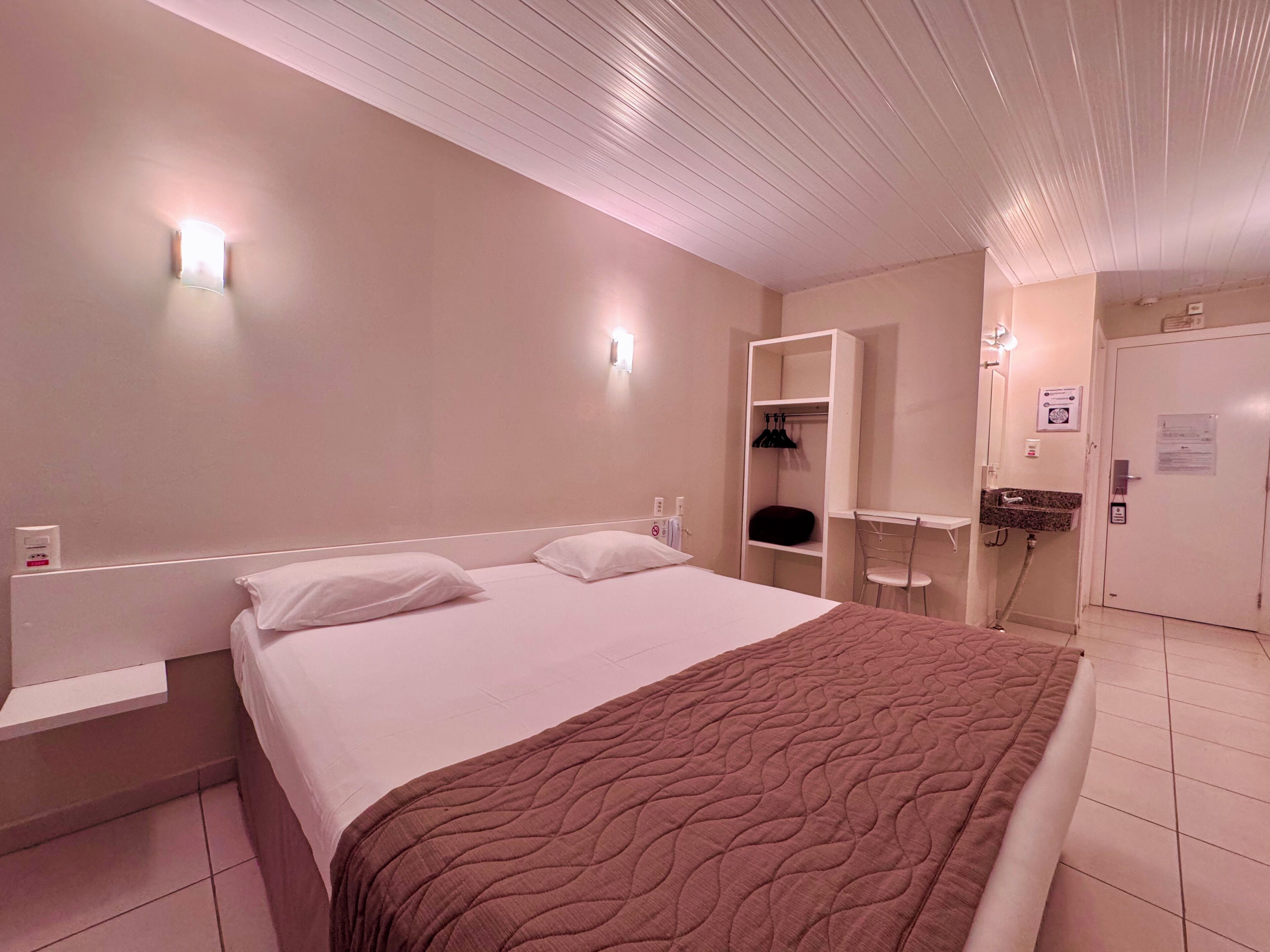 A hotel room with a bed, a nightstand, a wardrobe, a mirror, and a bathroom with a sink and a shower.
