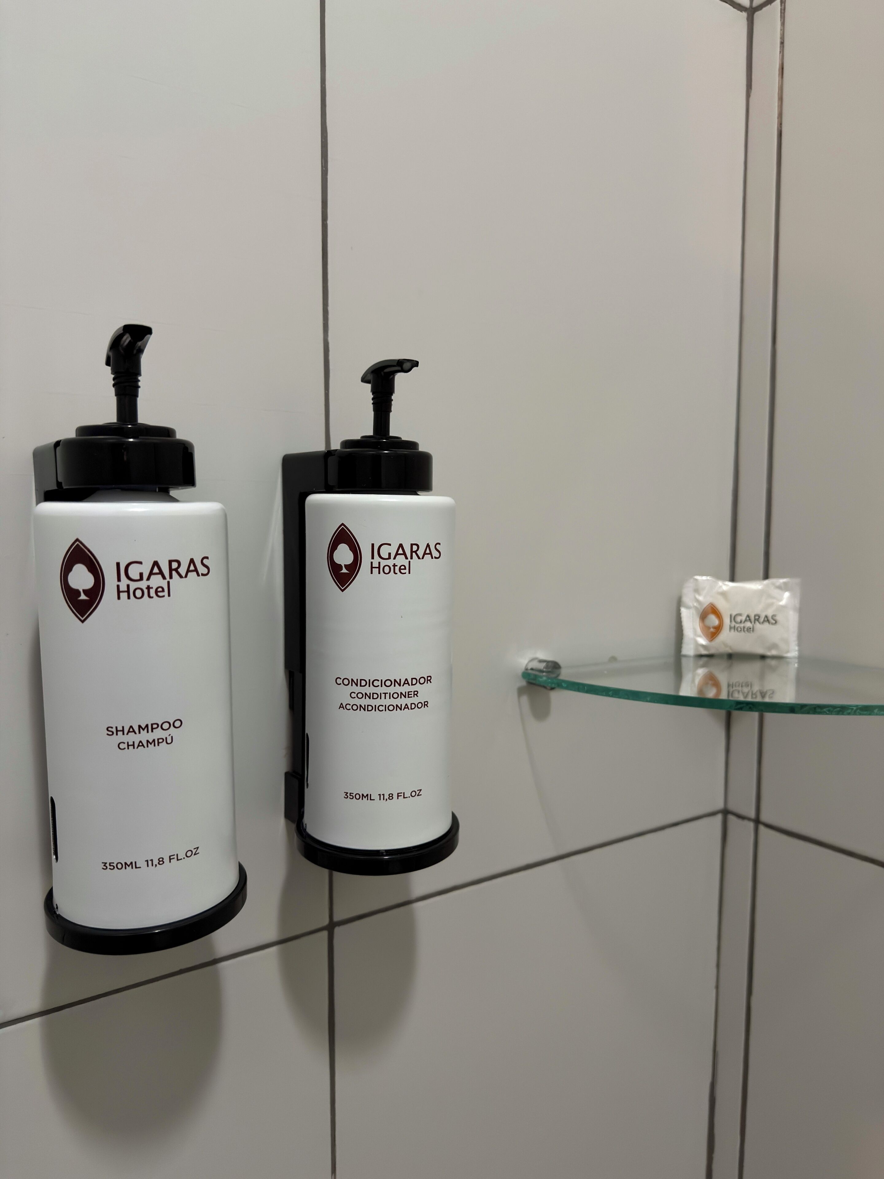 Two white hotel shampoo and conditioner bottles with black pumps, and a small glass container on a tiled wall.