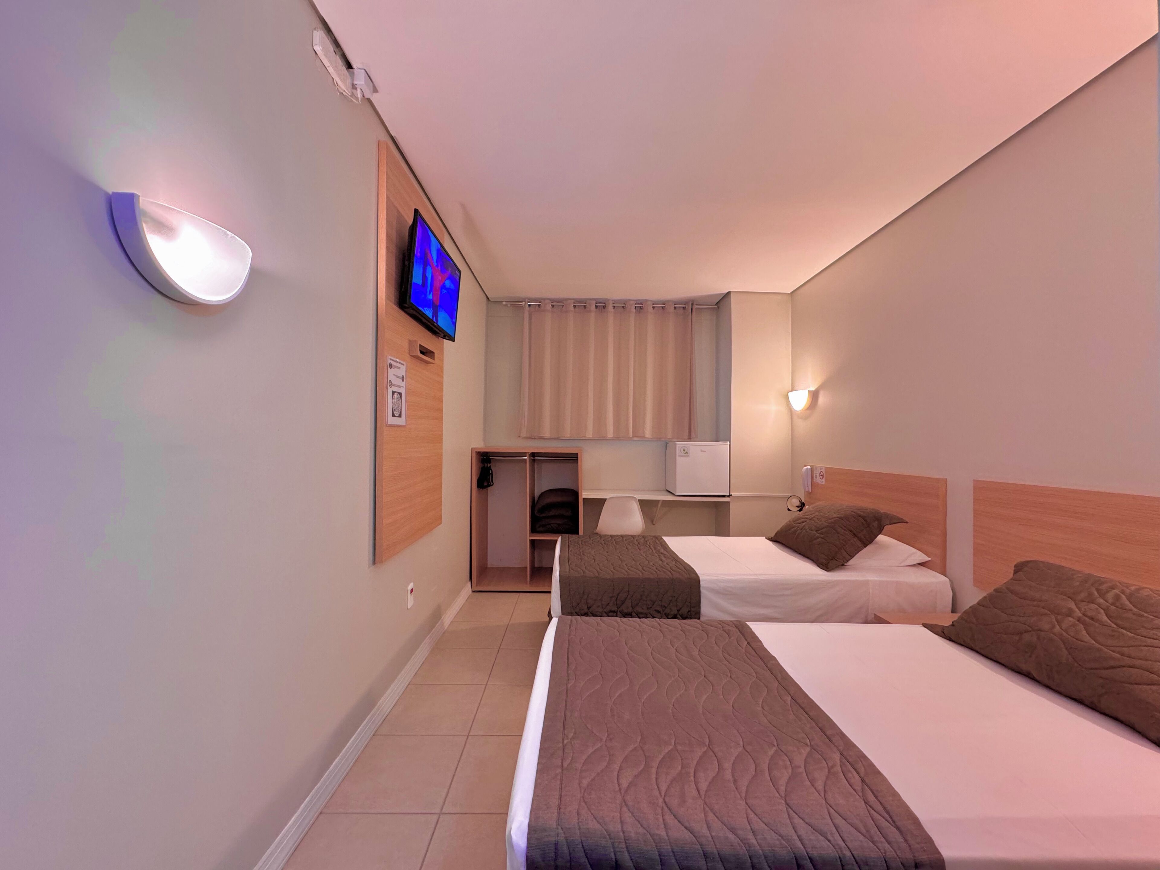 Habitación de hotel con dos camas, televisión, escritorio con silla, lámpara y ventana con cortinas.