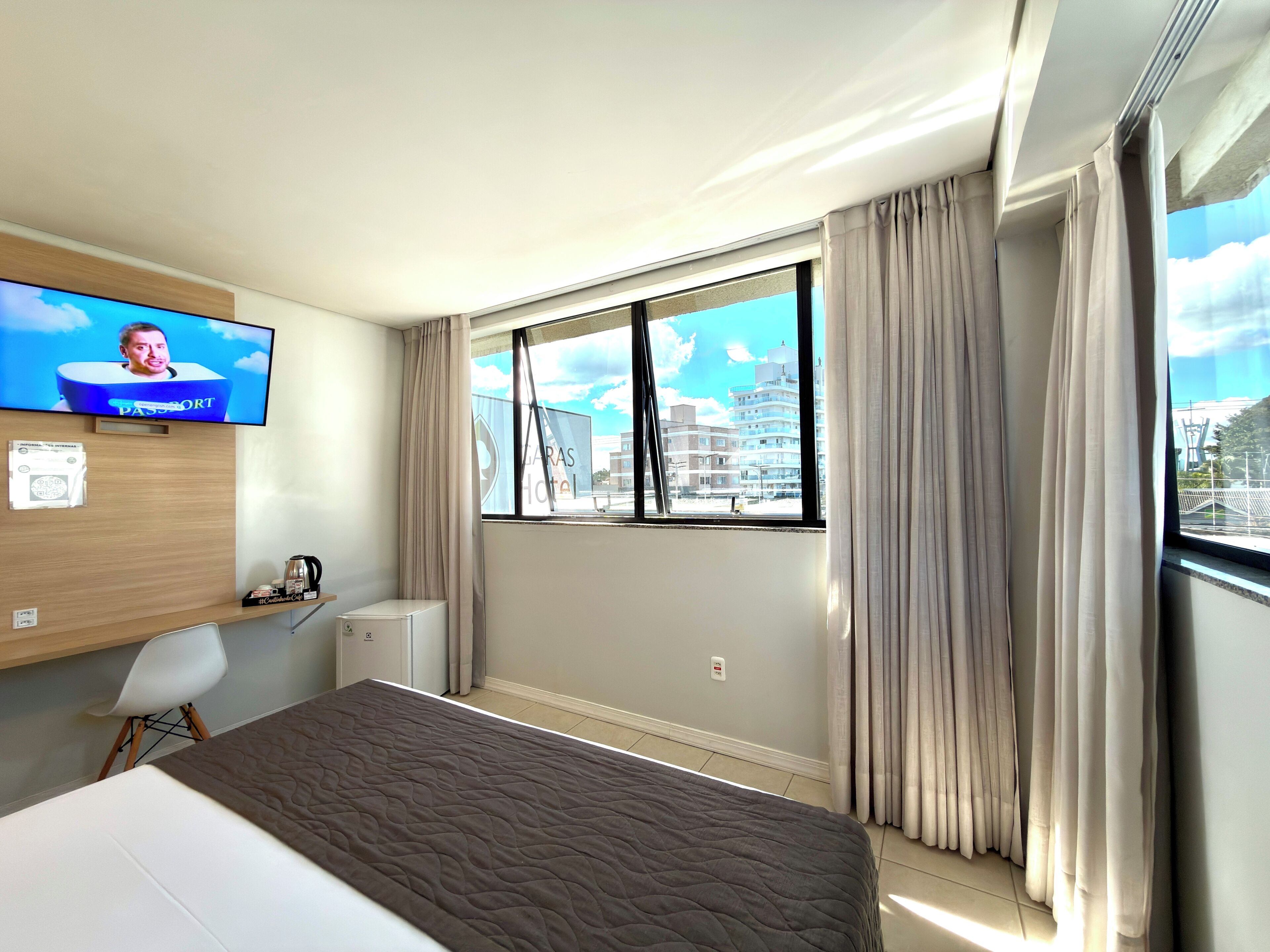 A hotel room with a bed, a television on the wall, a window with curtains, and a view of buildings outside.