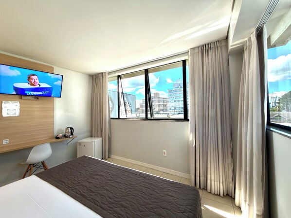 Habitación de hotel con cama, televisor en la pared, ventana con cortinas y vista a edificios.