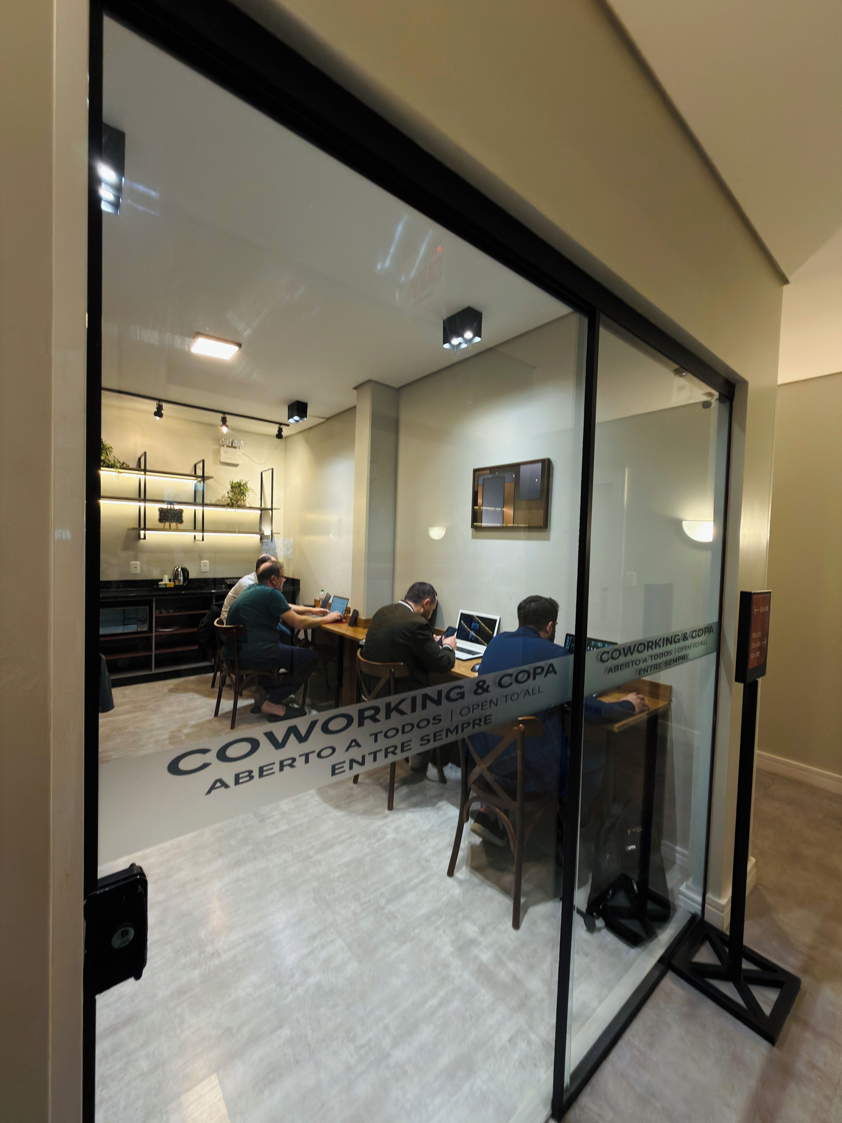 A modern office space with glass partitions, a kitchen area, and a sign indicating coworking and open to all.