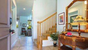 A hallway with a wooden staircase, a wicker console table with a lamp, and a large mirror. - Walk to Golf 250' from your door. Swim, Play, Feel the Warmth of Caring Owner (Waikoloa)