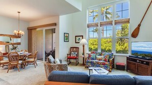 A spacious living room with a dining area, large windows with views of palm trees, a wooden ceiling, and a television. - Walk to Golf 250' from your door. Swim, Play, Feel the Warmth of Caring Owner (Waikoloa)