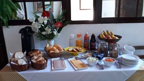 A breakfast buffet with bread, pastries, fruits, and beverages on a table by a window with curtains and framed pictures. - Casa das Orquídeas (Arraial do Cabo)