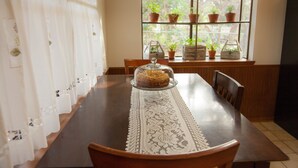 Un tavolo da pranzo con un runner in pizzo, un vassoio di vetro per dolci con una torta coperta e piante in vaso su uno scaffale vicino alla finestra.