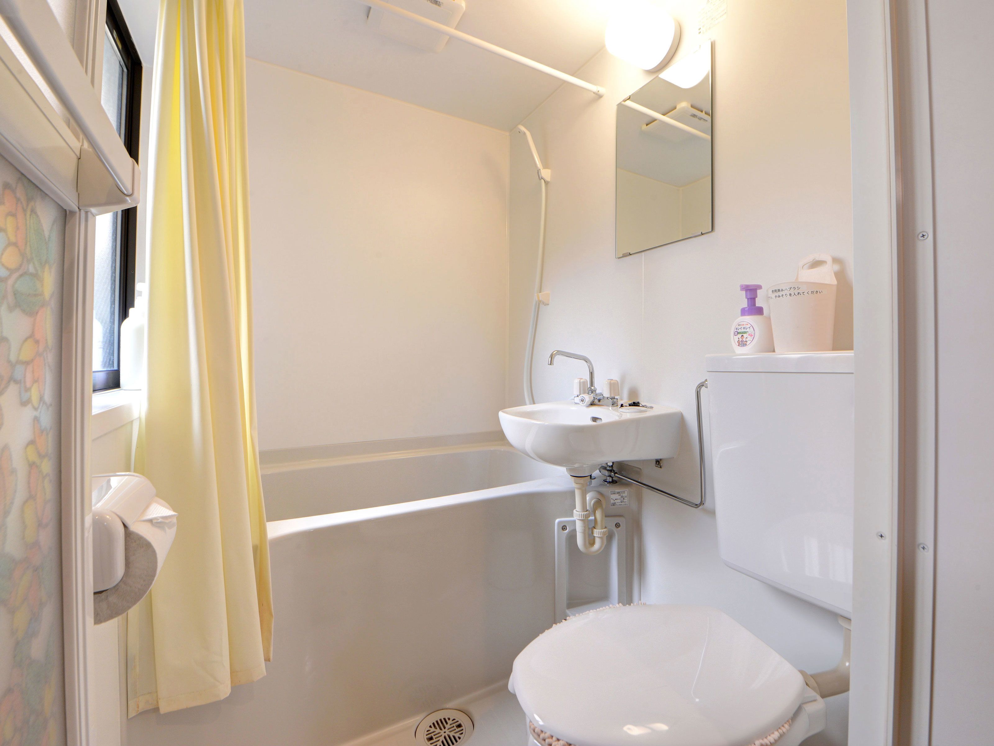 A bathroom with a white sink, toilet, and shower. There is a window with a curtain, and a mirror on the wall.