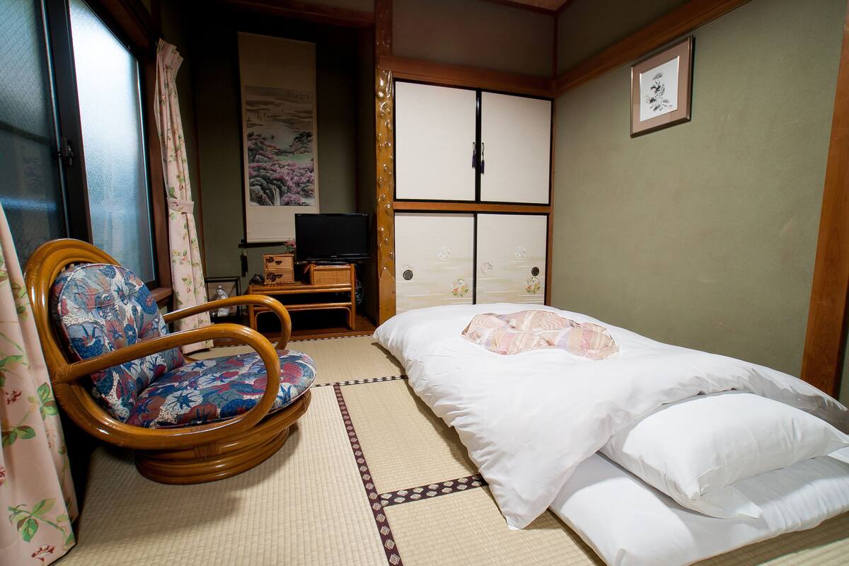 A traditional Japanese-style room with tatami flooring, a futon bed, a wooden rocking chair, and a television on a low shelf.