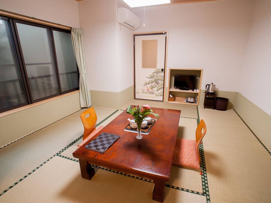 A traditional Japanese room with tatami flooring, a wooden table with a vase of flowers, a television, and a sliding door with a painting.