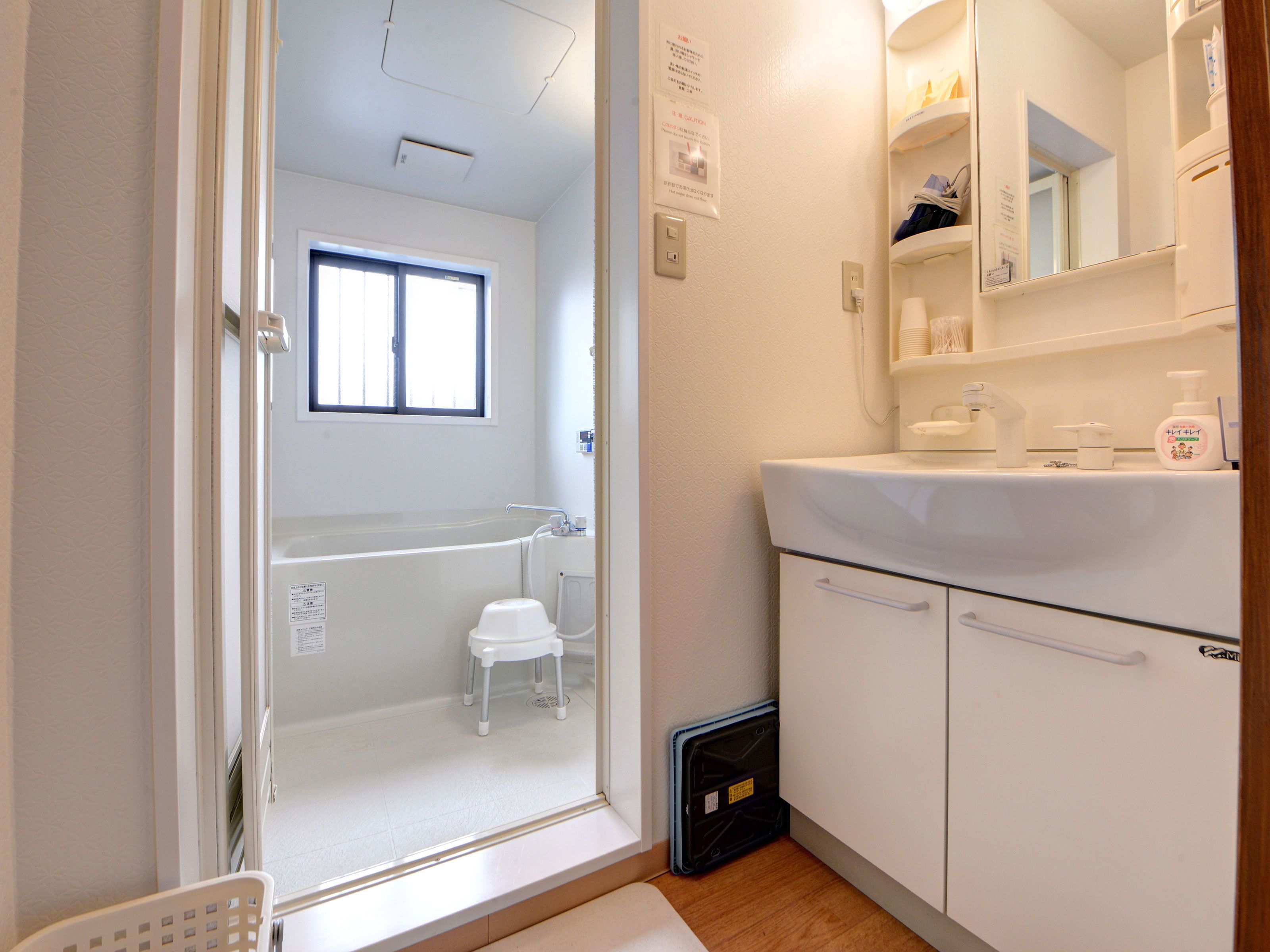 A bathroom with a bathtub, a sink, and a mirror. There is a window above the bathtub and a small stool near it.