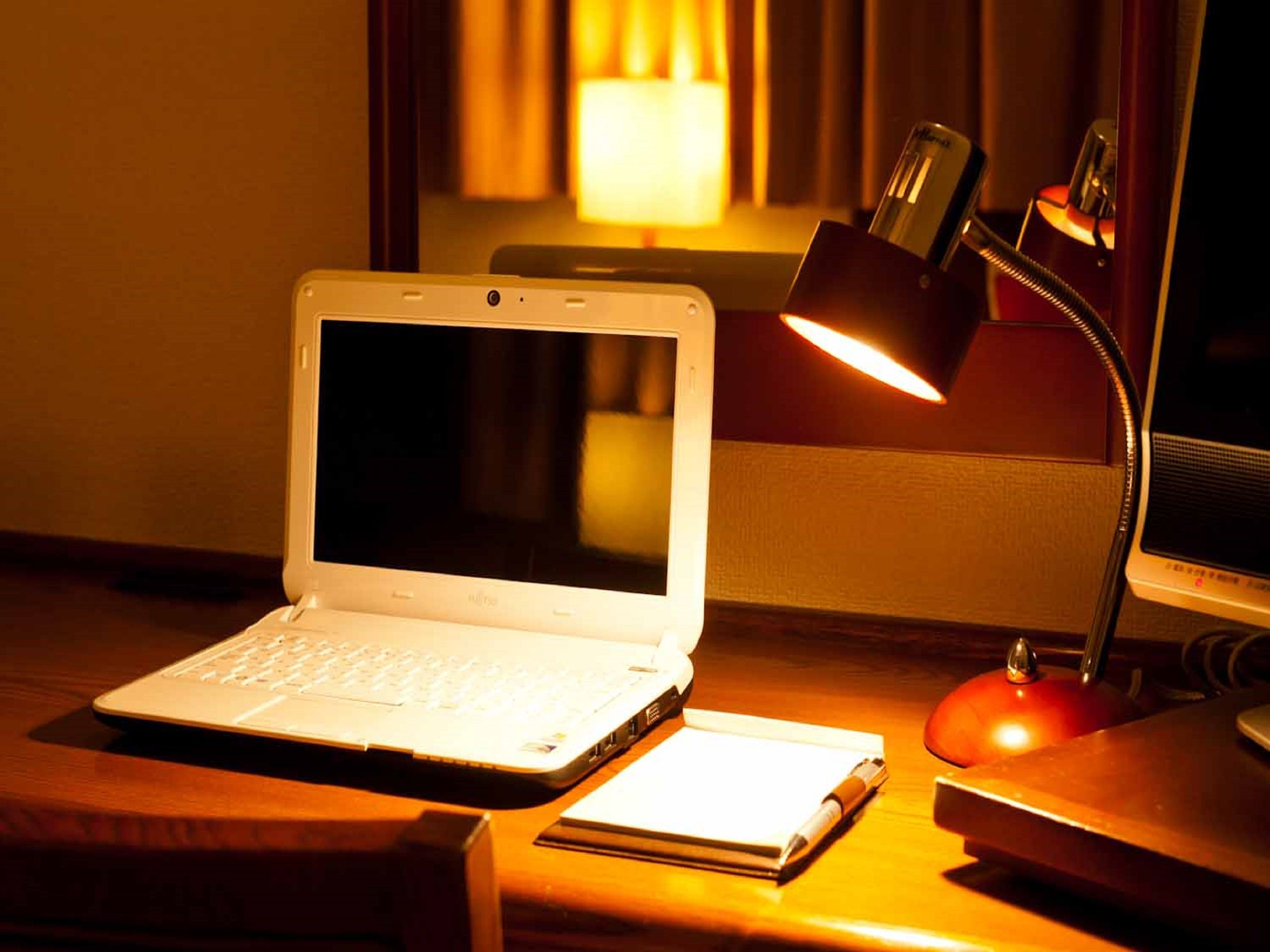 A laptop on a wooden desk with a notebook, a lamp, and a red object, possibly a speaker, in a warmly lit room.